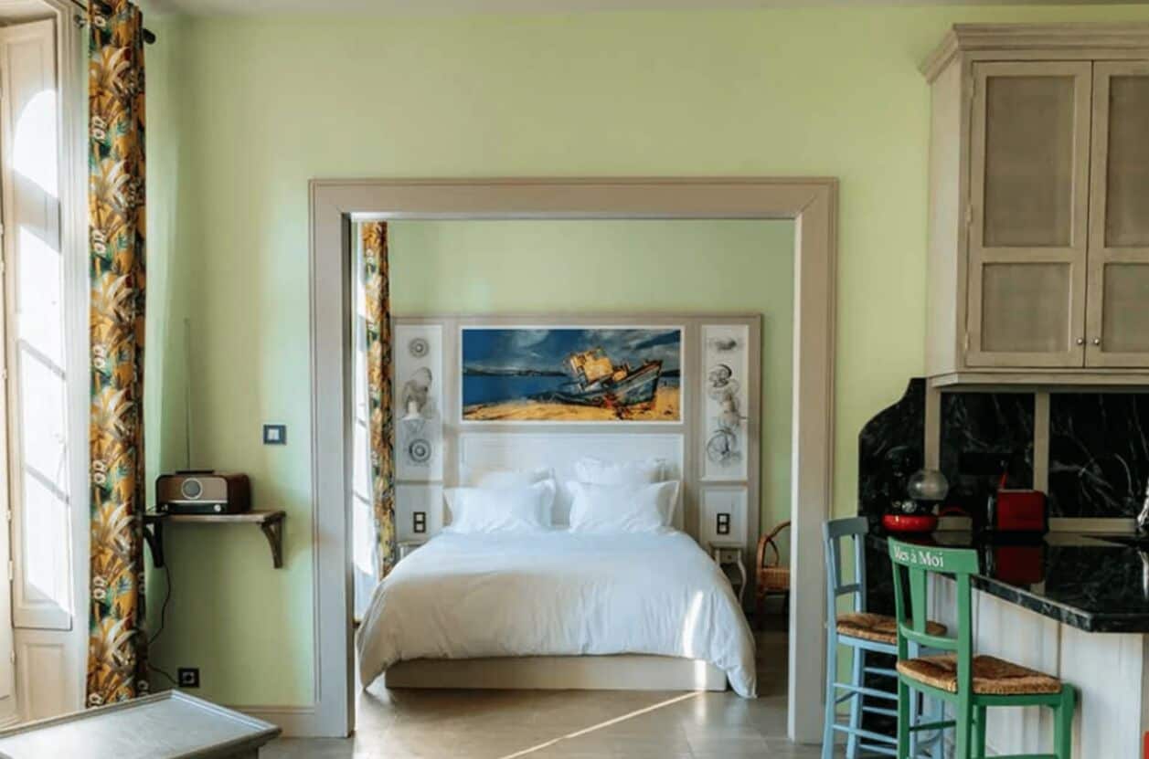 Un dormitorio con una cama, un cuadro en la cabecera, una mesita y una kitchenette con un taburete.