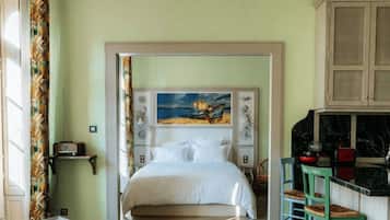 Un dormitorio con una cama, un cuadro en la cabecera, una mesita y una kitchenette con un taburete.