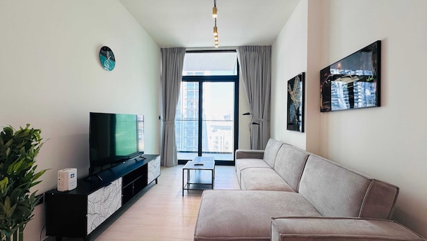 Una sala moderna con una televisión de pantalla plana sobre un soporte negro, un sofá color beige, una mesita auxiliar y un ventanal con cortinas.