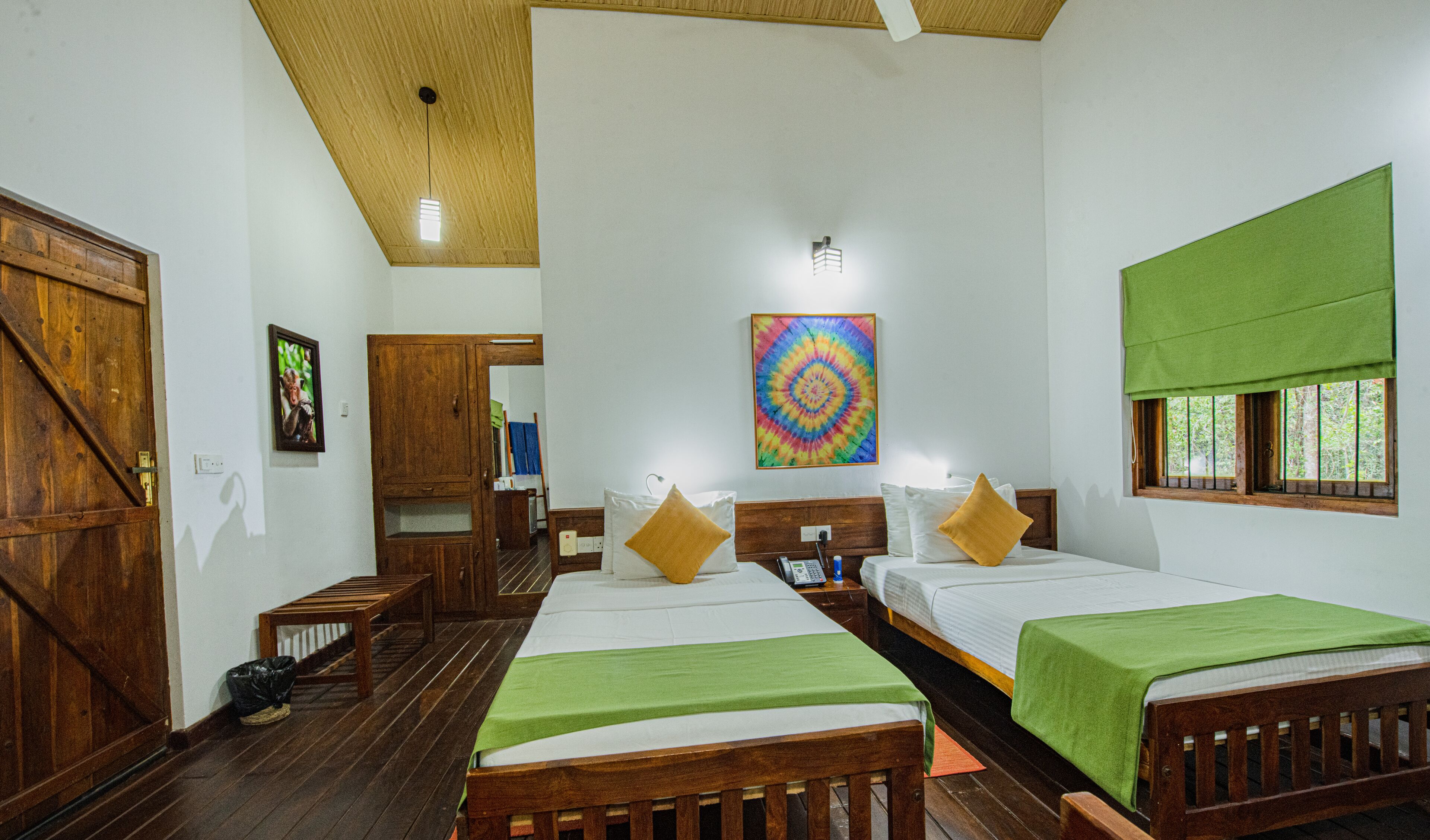 A room with two beds, wooden flooring, a wooden door, a green window blind, and a colorful wall art piece.