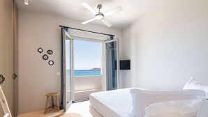 Een slaapkamer met een bed, een plafondventilator, uitzicht op zee en een klein krukje.