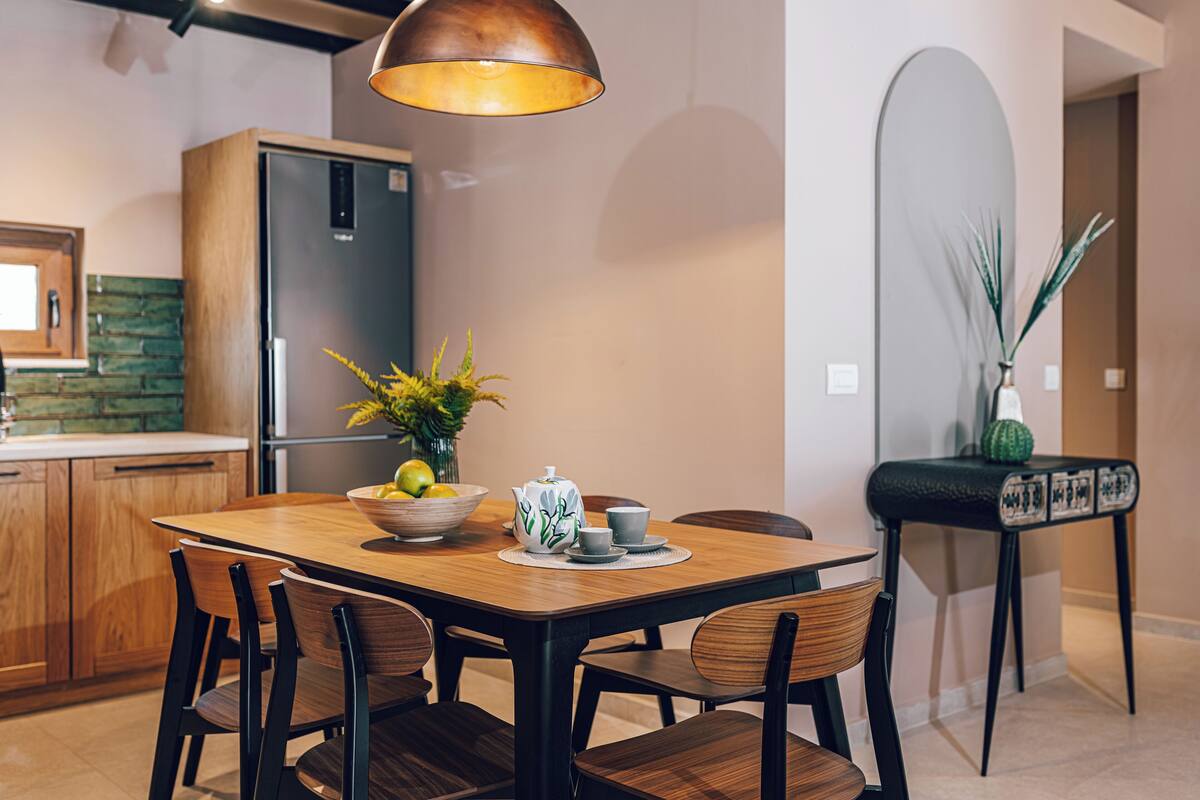 A modern kitchen with a wooden dining table, chairs, a refrigerator, and a hanging light fixture.