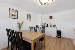 Un coin repas avec une table en bois, des chaises noires, un meuble blanc et une poubelle.
