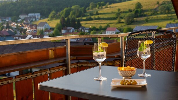 Un balcon avec une table, deux verres contenant une boisson claire agrémentée de rondelles de citron, un bol de collations et une vue panoramique sur un village.