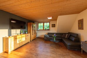 Een woonkamer met een houten plafond, een zwarte bank, een houten kast en een flatscreen televisie.