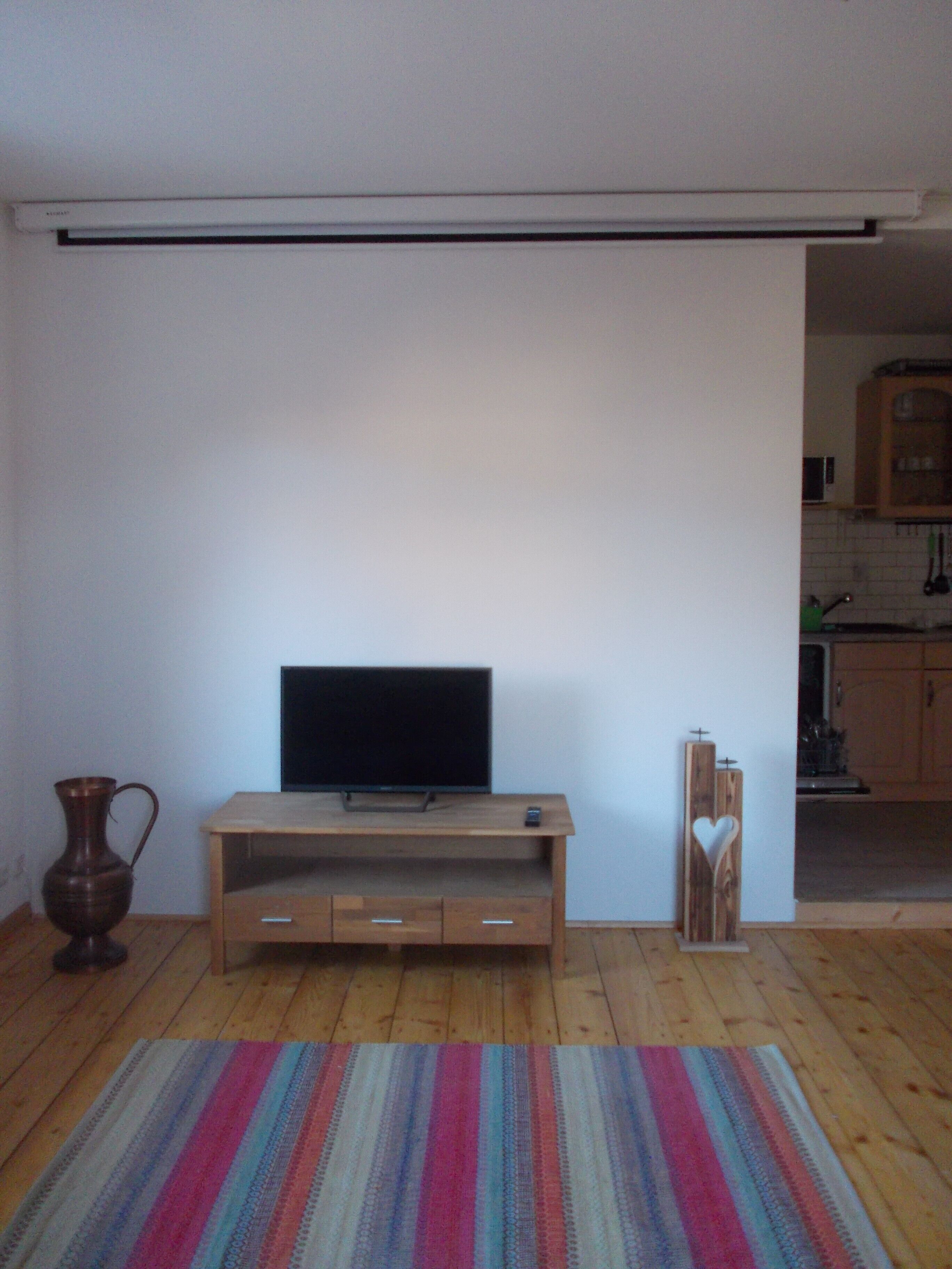 Une pièce avec un meuble TV en bois, un téléviseur à écran plat, un tapis à rayures, un vase et une décoration en forme de cœur.