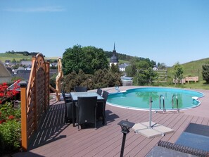 Ein Poolbereich mit Holzterrasse, Gartenmöbeln und Blick auf einen Hügel mit einer Kirche.