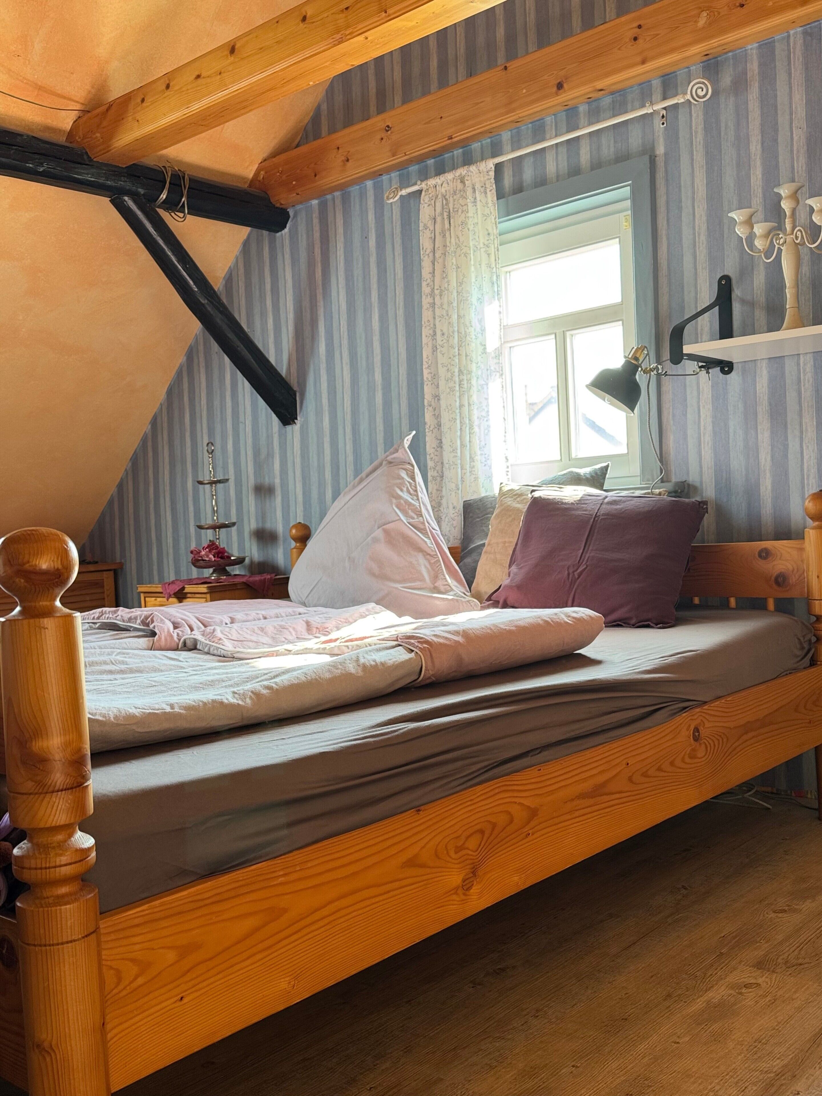 Ein hölzernes Bett in einem Raum mit gestreifter Tapete, ein Fenster mit Vorhängen und eine Schreibtischlampe.