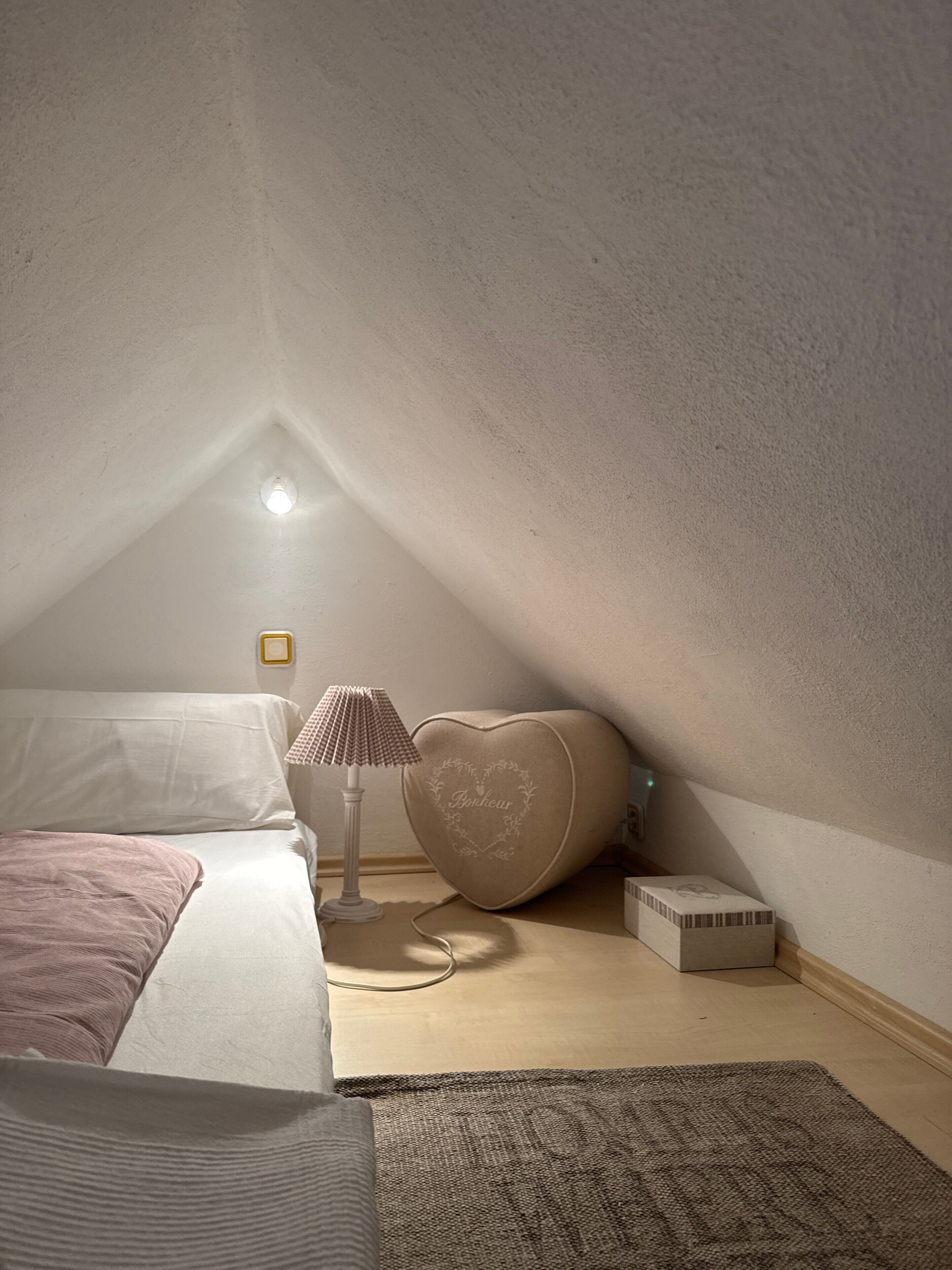 Quarto com teto inclinado, uma cama, mesa de cabeceira em formato de coração, luminária e pequena mesa de cabeceira com objeto decorativo.