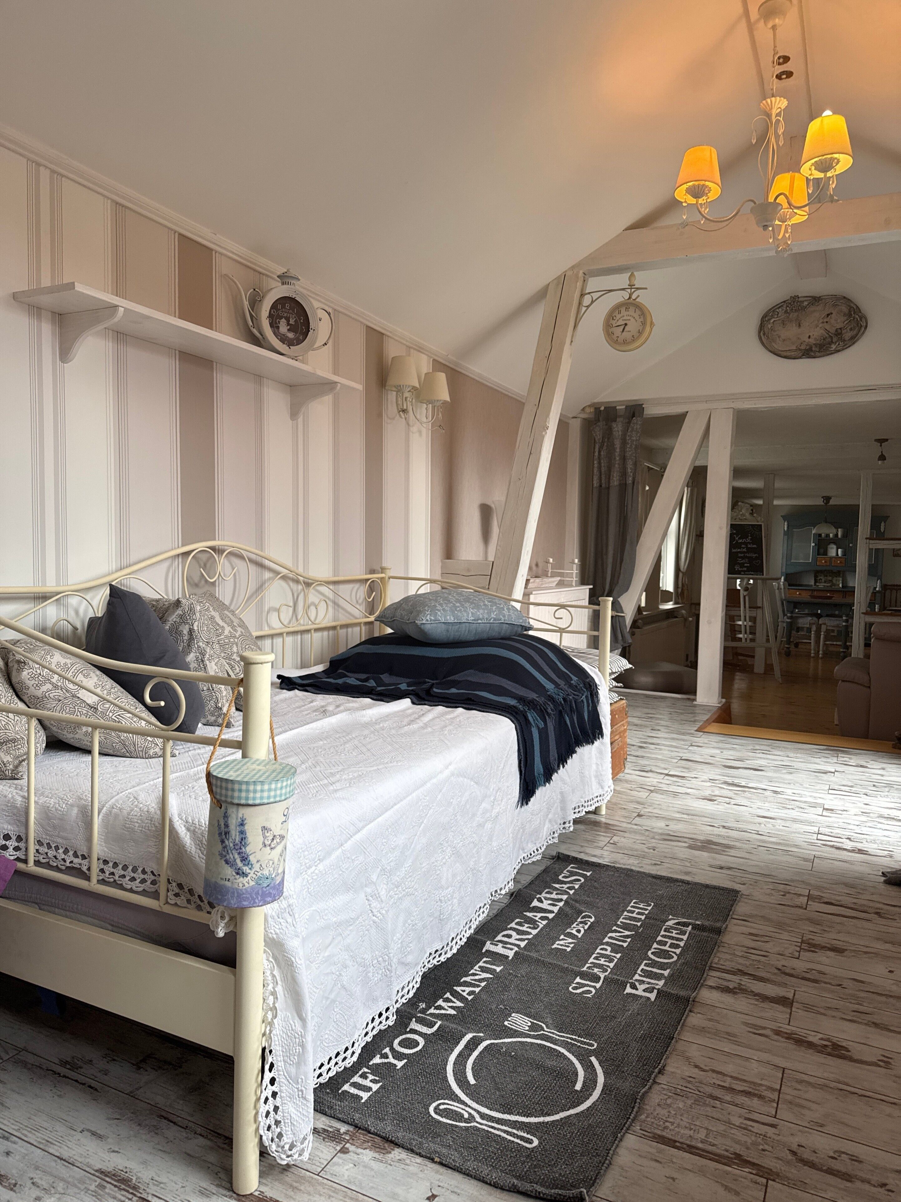 Een slaapkamer met een bed, nachtkastje, lamp, klok en uitzicht op de keuken.