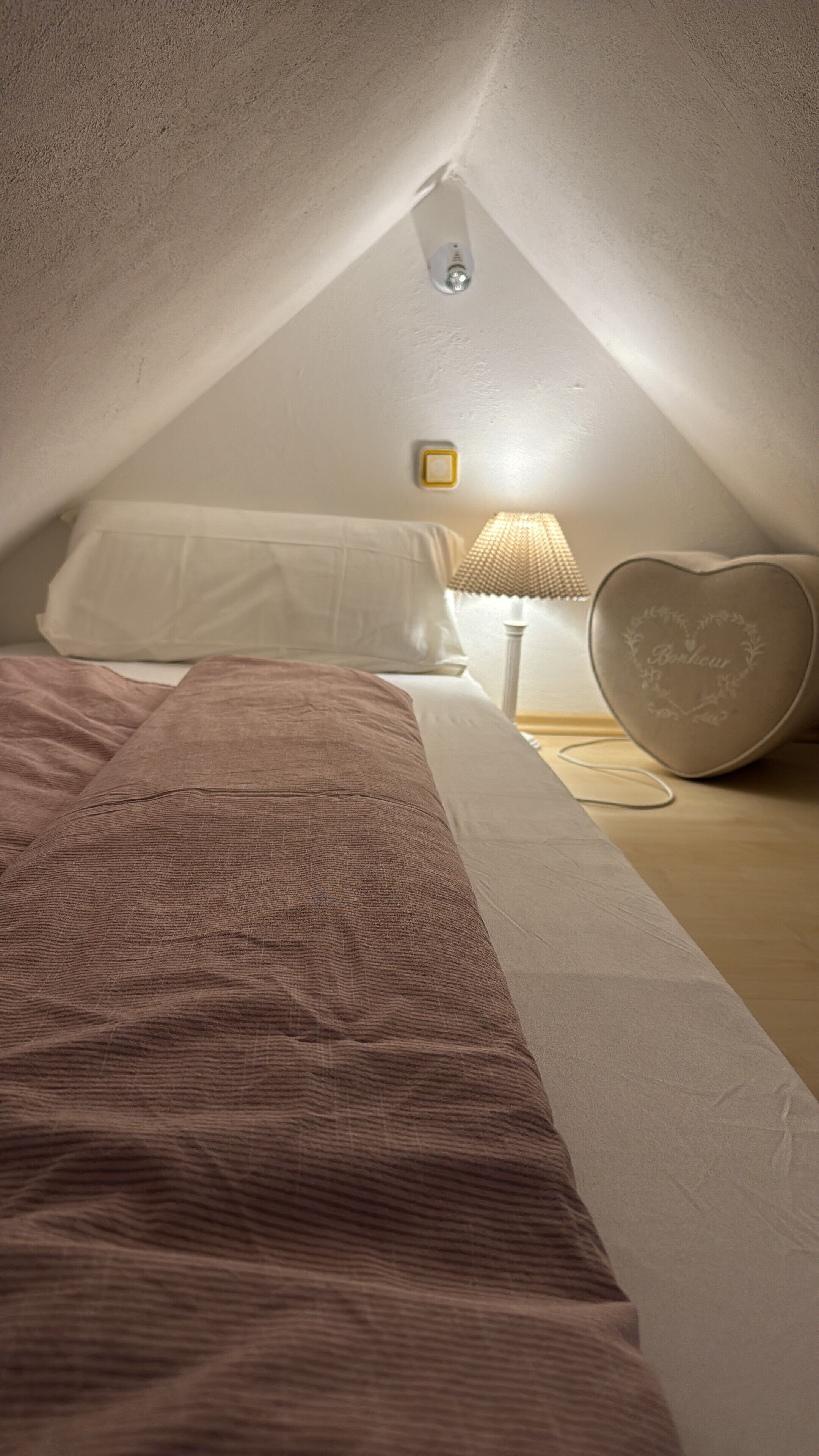 A cozy attic bedroom with a sloped ceiling, a bed with a pink blanket, a bedside lamp, and a heart-shaped decorative item on the nightstand.