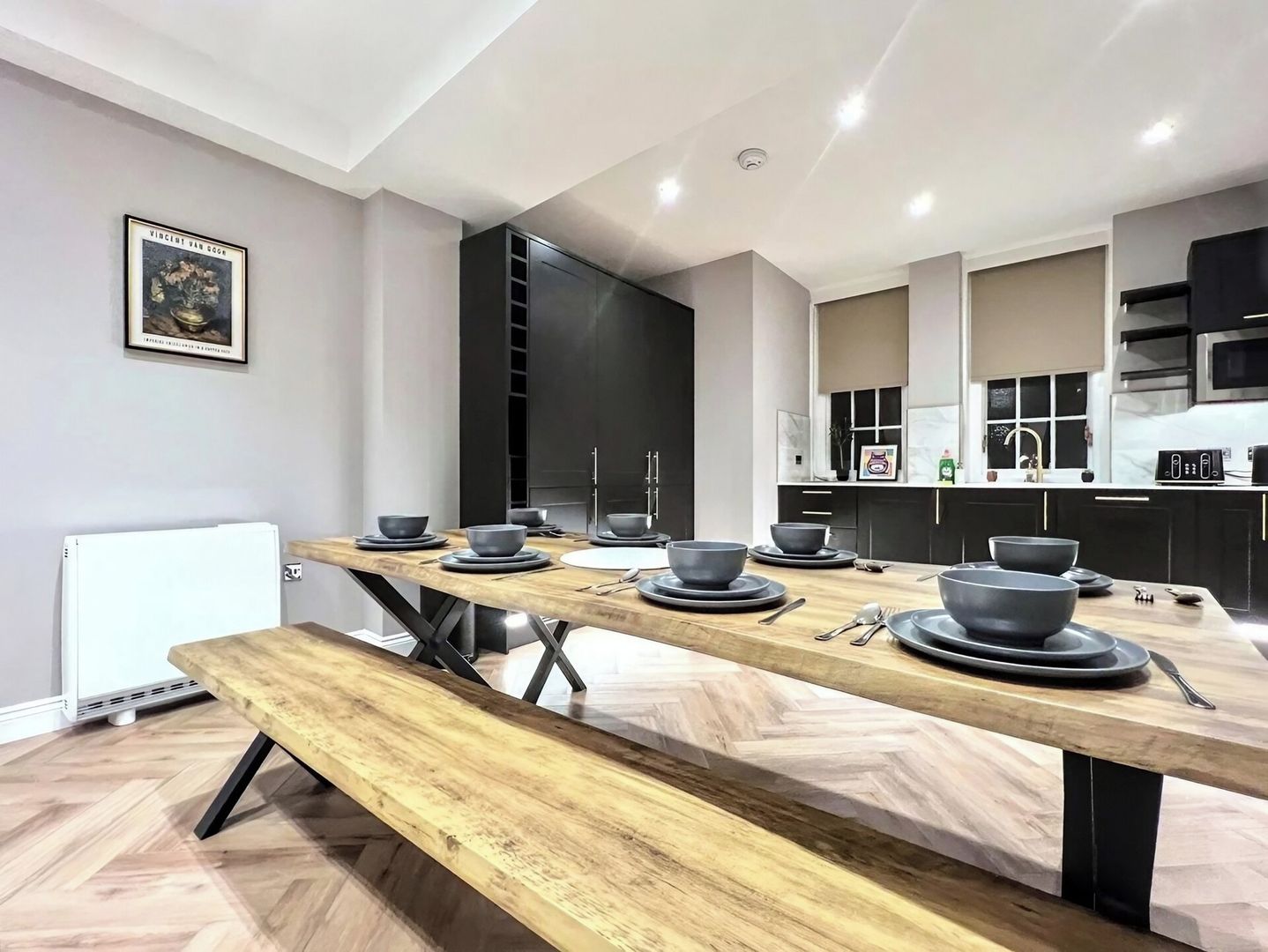 A modern kitchen with a dining area, featuring a long wooden table with grey bowls and plates, a bench, and a radiator.