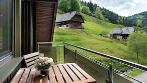 A balcony with a wooden table and chairs, a potted plant, and a view of a rural landscape with houses and hills. - Apartment 'Glashütte' with Mountain View, Balcony and Wi-Fi (Sankt Märgen)