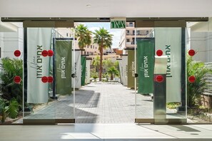 A hotel entrance with glass doors and green signage, a paved pathway, and palm trees in the background. - Red Canyon Spa Cozy Stay by Sea N' Rent (Eilat)