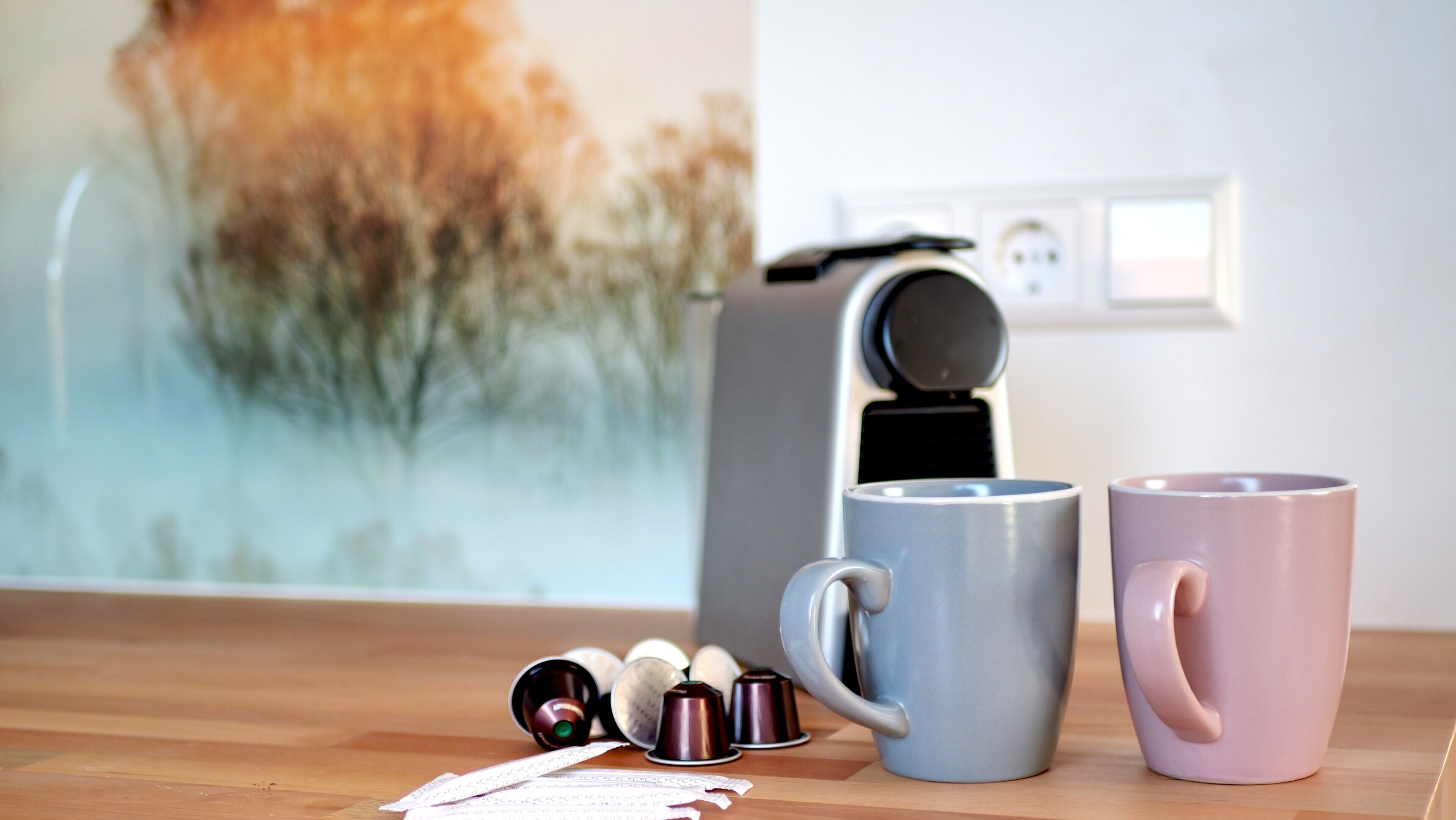 Une machine à café, deux tasses et des capsules de café sont posés sur une surface en bois, avec un tableau en arrière-plan.