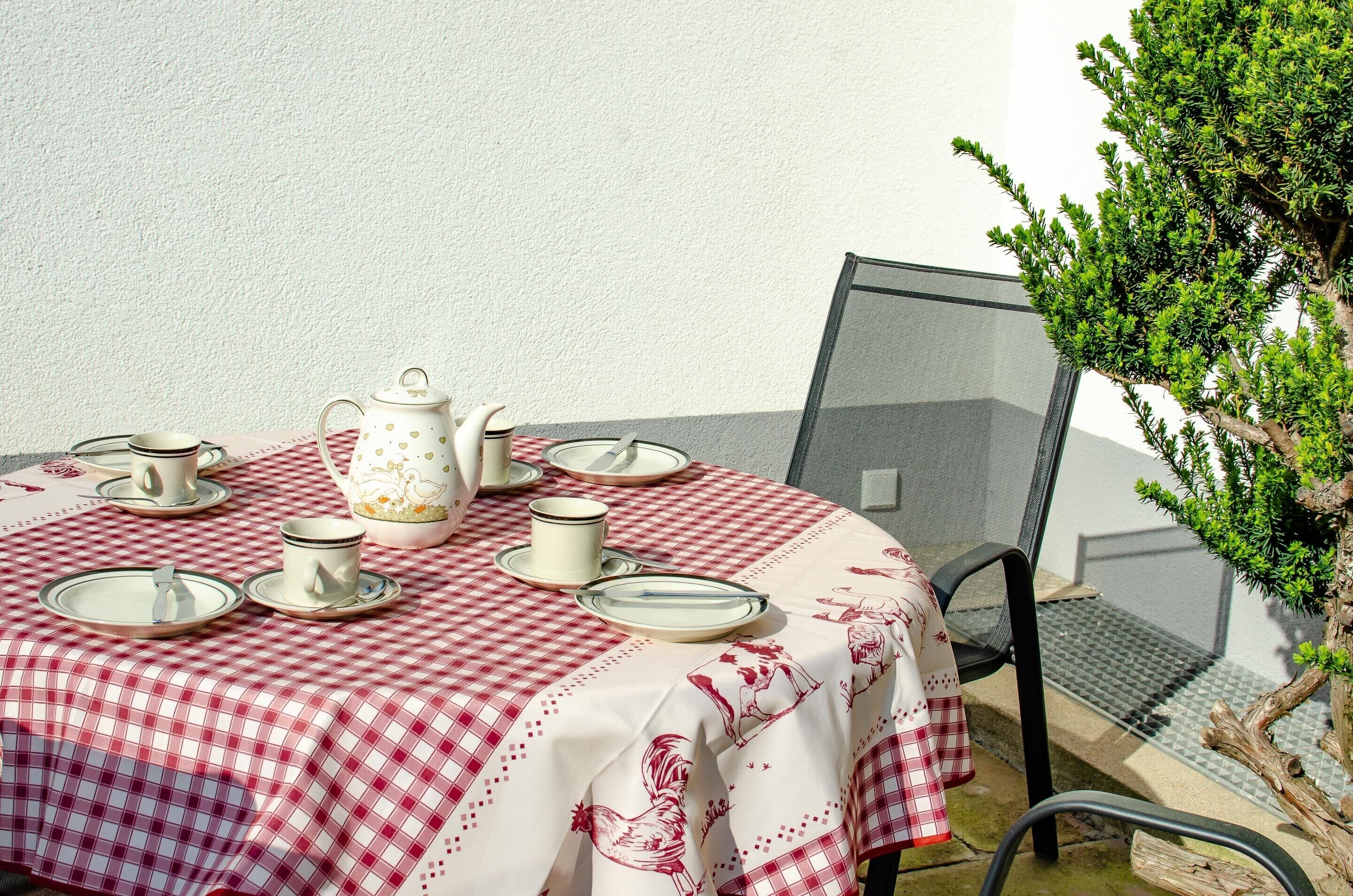 Una mesa servida con mantel de cuadros blancos y rojos, tazas de té blancas, platillos, una tetera y un vaso.