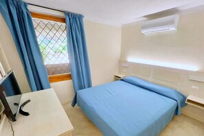 A hotel room with a bed, a desk with a computer, a window with curtains, and an air conditioning unit.