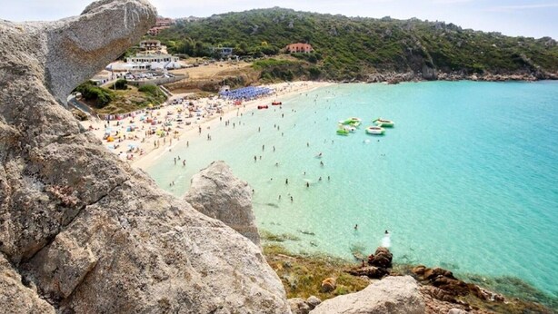 A coastal beach with clear turquoise waters, rocky cliffs, and a densely populated beach area with umbrellas and people. - Cosy apartment in Olbia (Olbia)