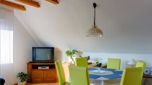 A dining area with a table set for four, a television on a wooden stand, and a hanging light fixture.