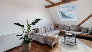 Ett rum i loftstil med synliga träbjälkar, en grå soffgrupp, ett runt soffbord och ett stort fönster med utsikt.