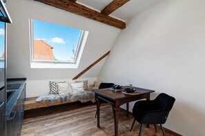 A loft-style room with a dining table, two chairs, a sofa, a window with a view of a red-roofed building, and exposed wooden beams. - Holiday Apartment 'Scheunenglück' with Balcony and Wi-Fi (Oberkirch)