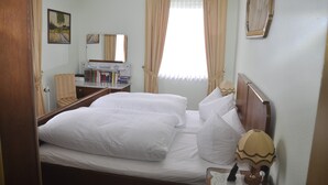 Habitación de hotel con dos camas, un escritorio con lámpara, una silla, una estantería y una ventana con cortinas.