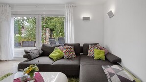 Uma sala de estar moderna com um sofá cinzento, almofadas coloridas, uma mesa de centro de vidro e vista através de uma porta de vidro.