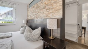 Quarto moderno com parede de tijolos decorativa, cama com almofadas estampadas, mesa de cabeceira com abajur e janela com cortinas translúcidas.