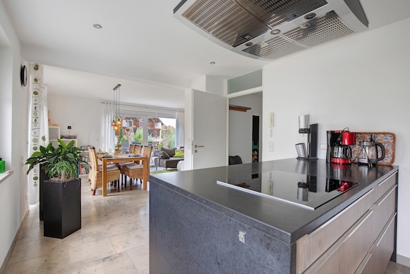 Une cuisine moderne dotée d’un îlot central, d’un coin repas avec une table et des chaises, et d’une vue sur un balcon accessible par une porte ouverte.