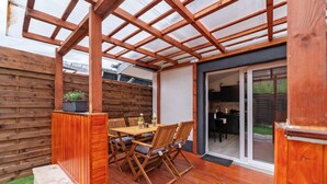 A covered patio with a wooden table and chairs, a wooden fence, and a sliding glass door leading to an indoor area. - Holiday Home 'Zum Stumpfen Turm' with Sauna, Private Terrace and Garden (Morbach)