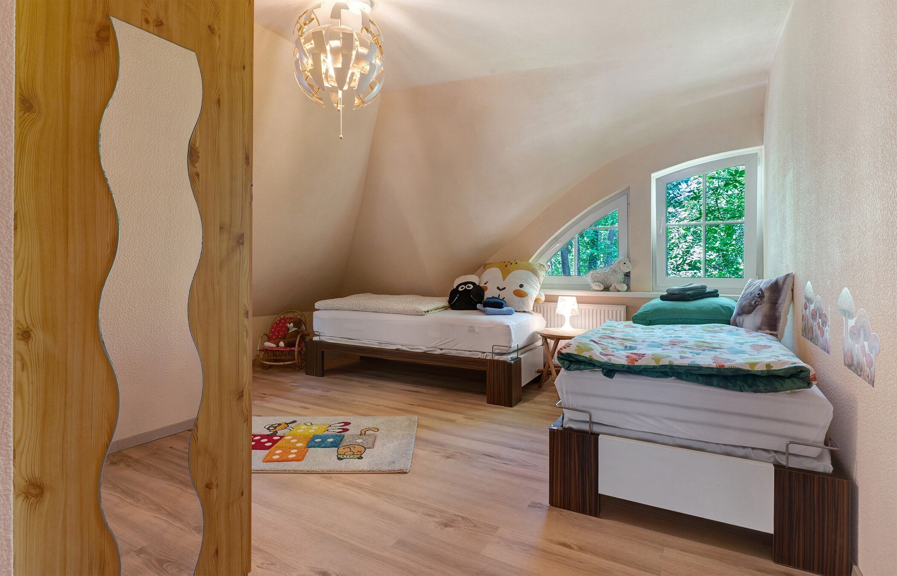 Ein Schlafzimmer mit schrägem Dach, zwei Betten, Holzboden, einer Kronleuchter, einem Fenster mit Blick auf Bäume und einem kleinen Tisch mit Lampe.