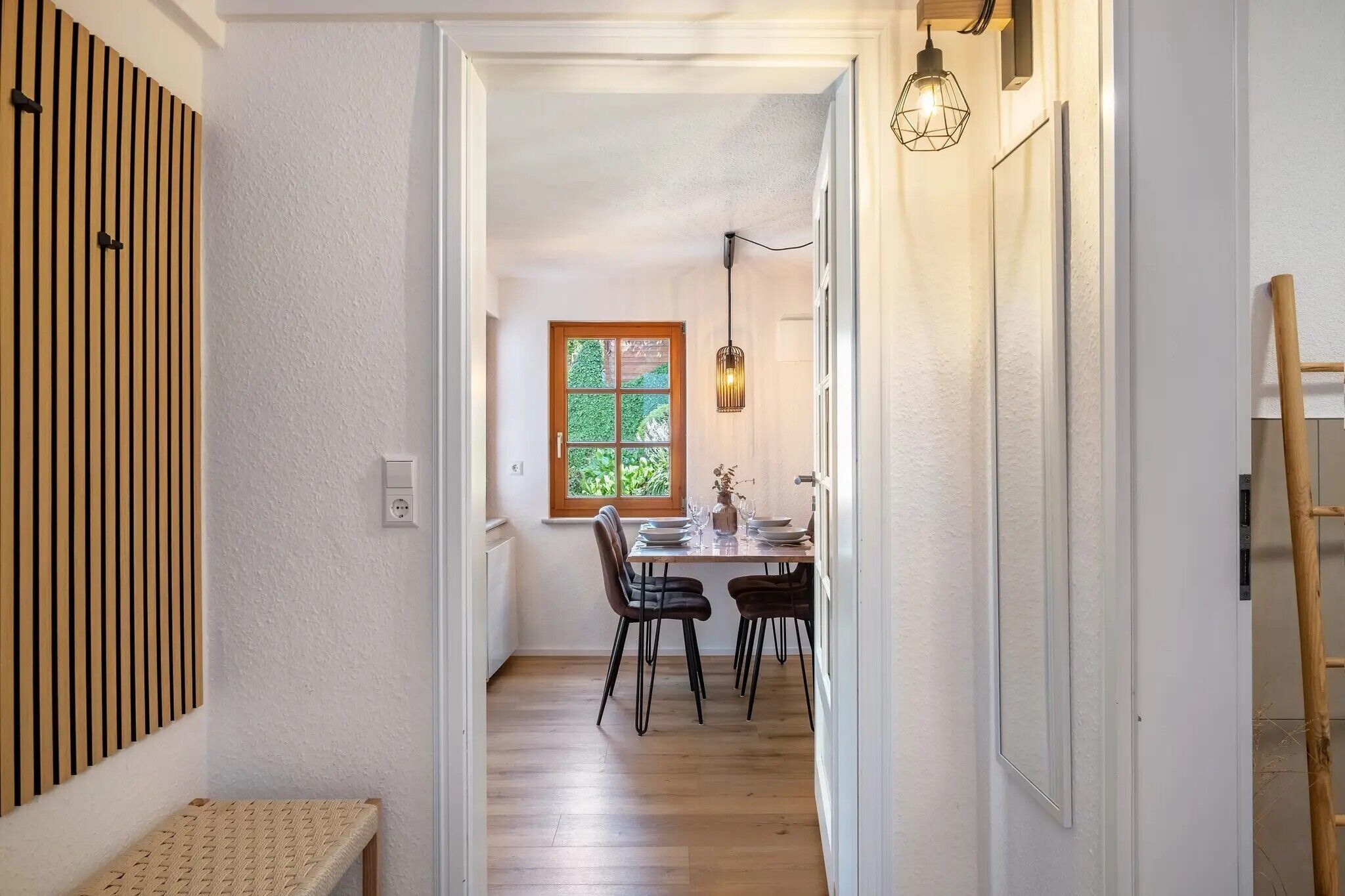 Un corridoio che conduce a una sala da pranzo con una panca di legno, un tavolo apparecchiato per due e una finestra con vista sul verde.