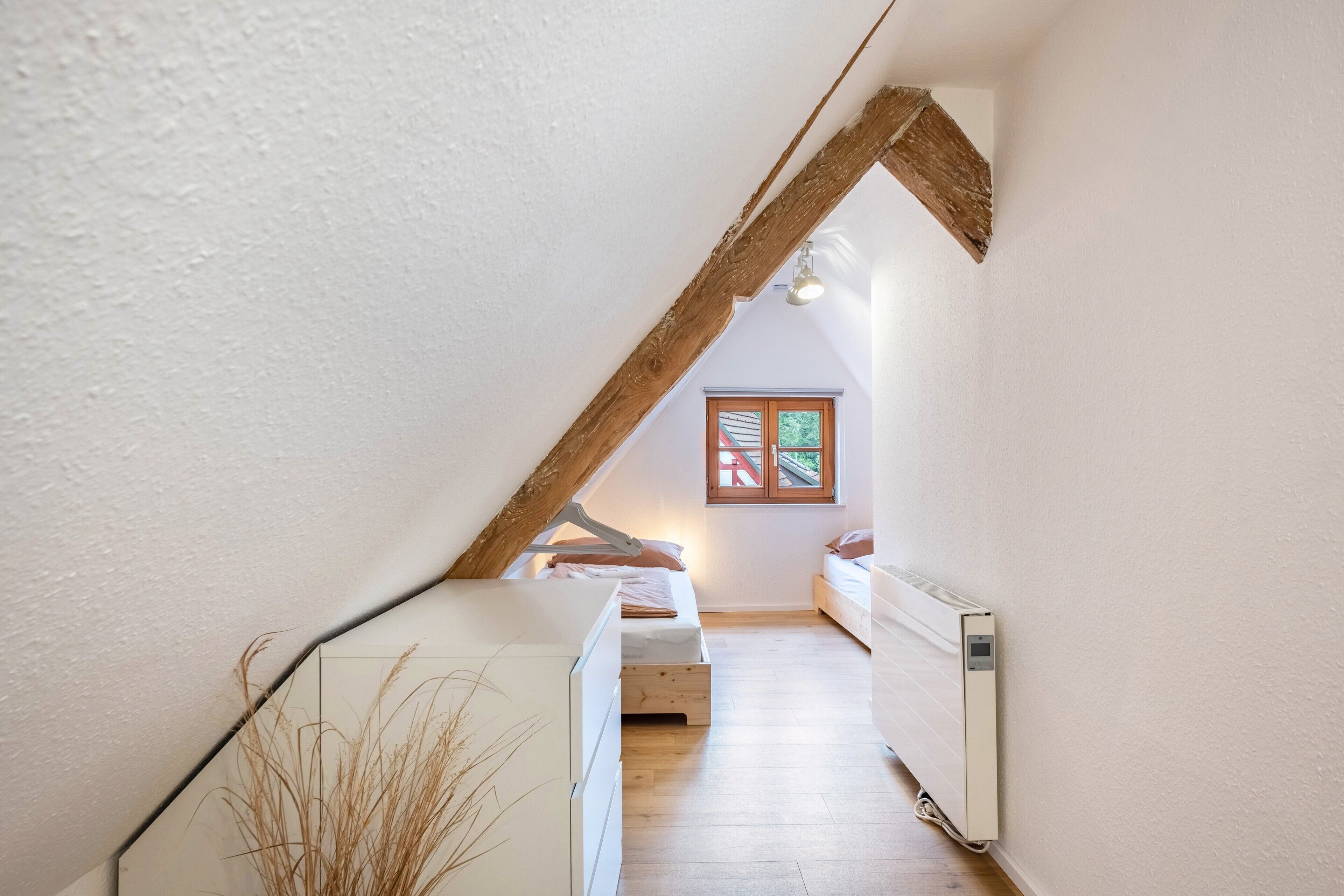 Ein schmales Schlafzimmer im Dachgeschoss mit schrägen Wänden, Holzbalken, einer Heizung und einem Fenster mit Blick.