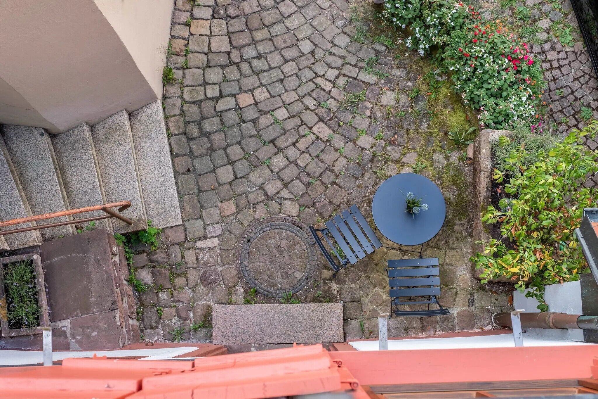 Une cour avec un chemin pavé de pierres, une table ronde entourée de chaises et un petit jardin fleuri.