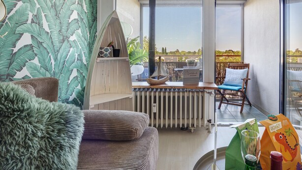 A modern living room with a view of the city, a cozy sofa, a wooden side table, and a chair with a cushion. - Apartment 'Bootsmann Steinhude' with Private Terrace, Balcony and Wi-Fi (Wunstorf)