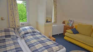 A bedroom with two beds, a window with curtains, a radiator, a wooden nightstand, and a yellow sofa.