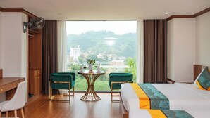 A hotel room with a bed, a desk, two chairs, a small table with a vase, and a view of a cityscape and hills.
