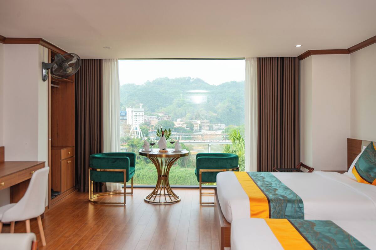 A hotel room with a bed, a desk, two chairs, a small table with a vase, and a view of a cityscape and hills.