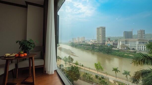 A room with a large window overlooking a river, a table with fruits and a plant, and a view of the city skyline.