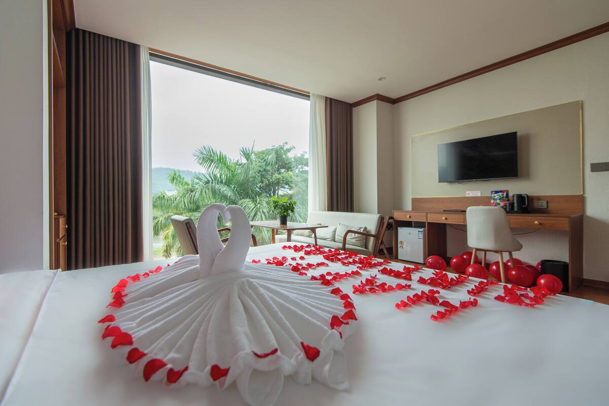 A hotel room with a bed adorned with a swan-shaped towel and red rose petals, a desk with a computer, a chair, and a television on the wall.