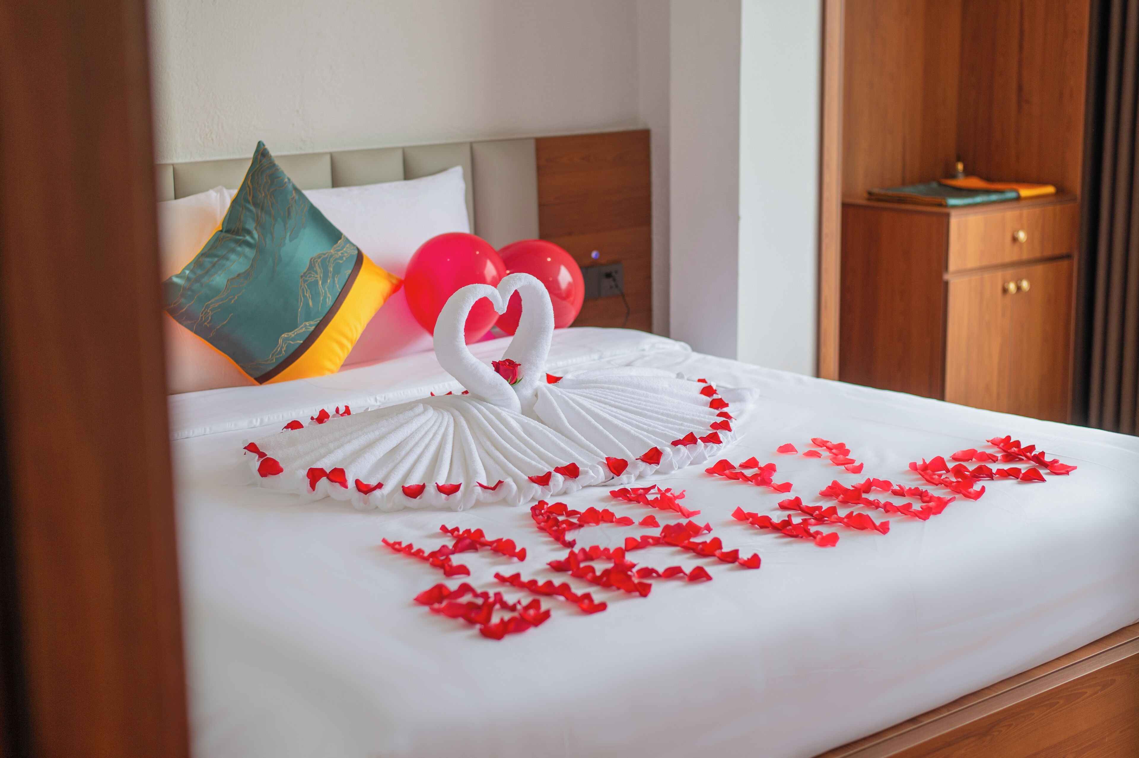 A neatly made bed with a heart-shaped towel arrangement and red rose petals.