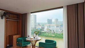 A hotel room with a large window, a green sofa, a small round table with a vase, and a bed with a yellow and green bedspread.