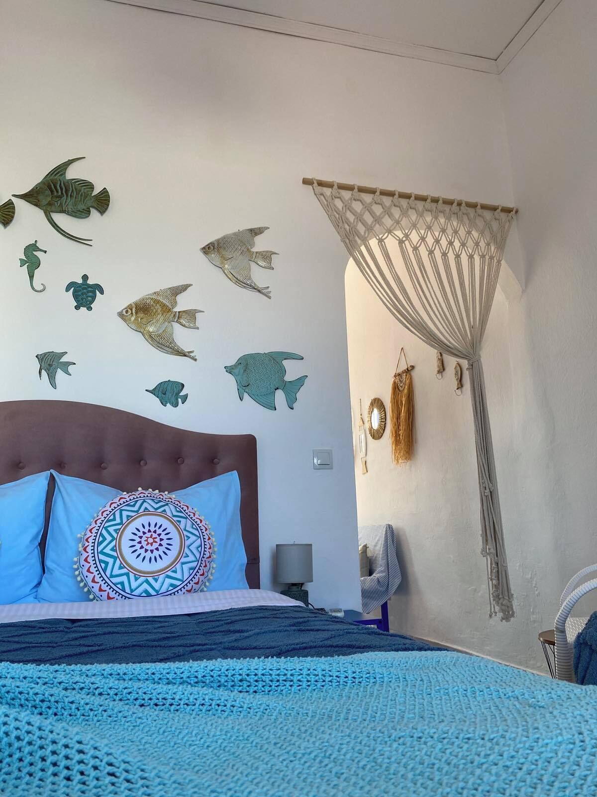 A bedroom with a bed, a wall decorated with fish and seahorse wall art, a macrame curtain, and a patterned throw blanket.