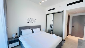 Una habitación de hotel moderna con una cama grande, una mesita de noche con lámpara, una obra de arte geométrica en la pared, un espejo grande y una puerta que da a otra habitación.