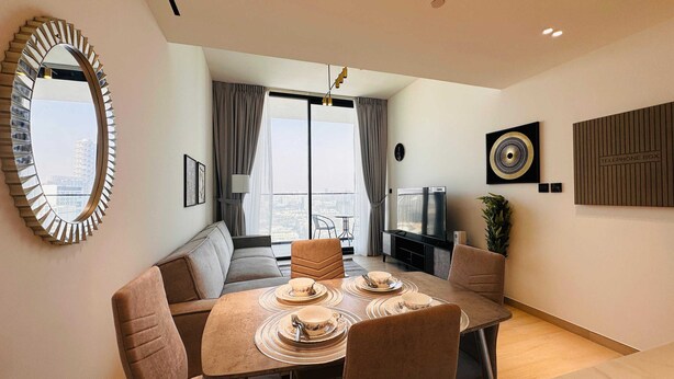 Una sala de estar moderna con un sofá, una mesa de comedor preparada para cuatro personas, un televisor y vistas a la ciudad a través de grandes ventanales.