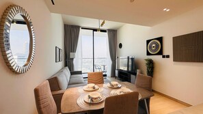 A modern living room with a sofa, dining table set for four, a television, and a city view through large windows. - OYO HOME 1483 One Bedroom Apartments  (Dubai)