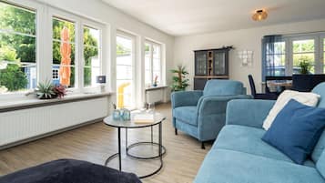 Ein modernes Wohnzimmer mit einem blauen Sofagarnitur, einem runden Couchtisch und Blick ins Freie durch große Fenster.