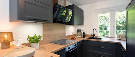 A modern kitchen with dark cabinetry, a wooden countertop, a built-in oven, a sink, and a window providing a view of greenery outside.