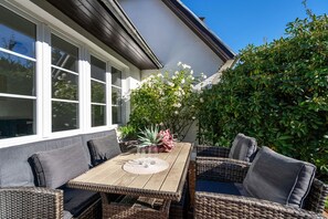 An outdoor dining area with a wooden table and wicker chairs, surrounded by greenery and a white building. - Nala's Hüs - Beautiful house for 6 people with a terrace, parking, and Wi-Fi. (Westerland)