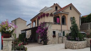 Una casa de dos pisos con fachada de piedra, un balcón adornado con flores moradas y un patio cubierto.
